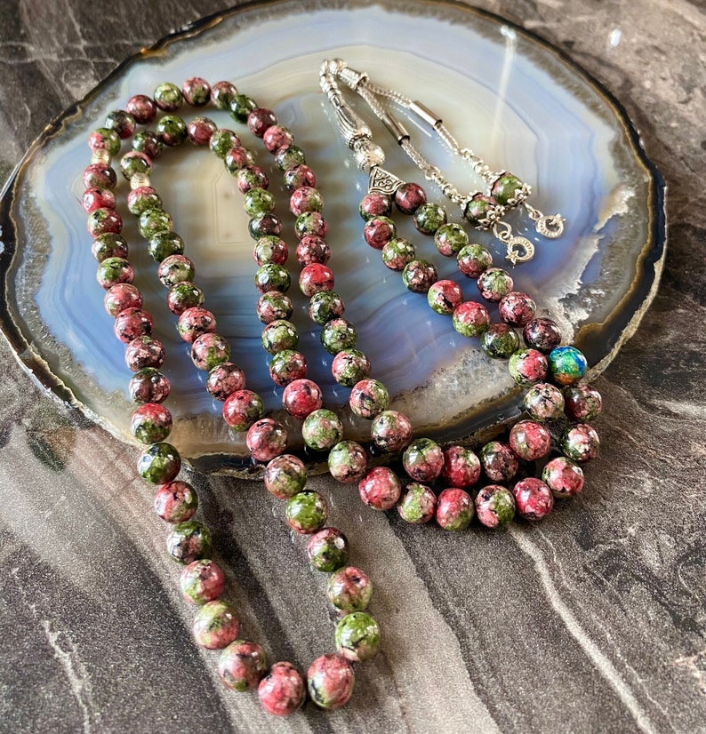 May include: A prayer bead mala made with green and pink speckled beads. The mala is laid out on a gray and white agate slab.