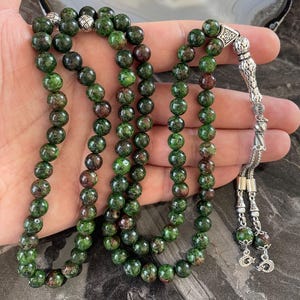 May include: A green and brown gemstone prayer beads mala with a silver tassel and a silver charm. The beads are round and have a speckled appearance.