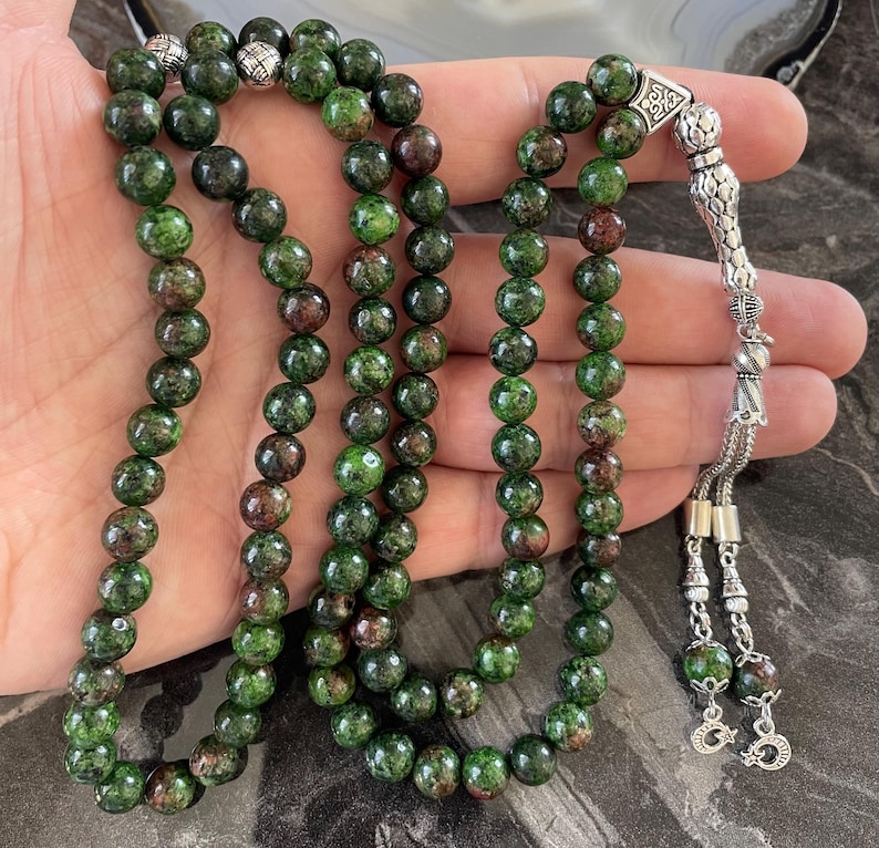 May include: A green and brown gemstone prayer beads mala with a silver tassel and a silver charm. The beads are round and have a speckled appearance.