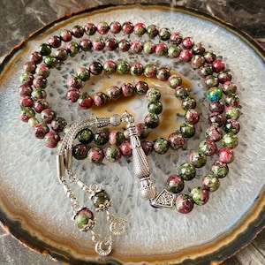 May include: A green and pink prayer beads mala with silver accents. The beads are made of a speckled stone with a green and pink pattern. The mala is laid out on a white and gray agate slice.
