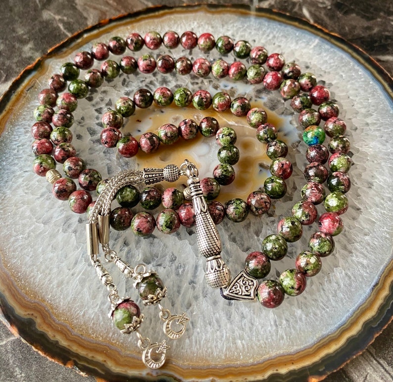 May include: A green and pink prayer beads mala with silver accents. The beads are made of a speckled stone with a green and pink pattern. The mala is laid out on a white and gray agate slice.