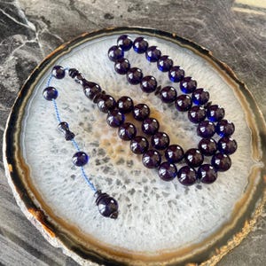 May include: A dark blue prayer bead set with a tassel and a blue string. The beads are round and polished, with a deep, reflective color. The set is displayed on a white and brown patterned surface.