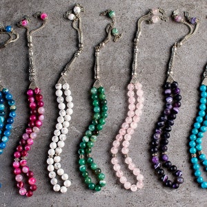 May include: Seven strands of colorful gemstone prayer beads with silver accents. The beads are blue, pink, white, green, pink, purple, and turquoise.