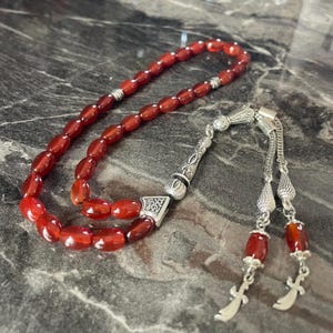 Può includere: Un rosario di preghiera rosso con dettagli argentati. Le perline ovali sono collegate da maglie argentate. Il rosario presenta un ciondolo decorativo argentato e due nappe argentate con gocce di perline rosse.