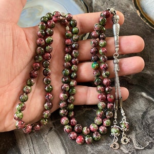 May include: A green and pink prayer beads mala with silver accents. The beads are round and have a speckled pattern. The mala is strung on a silver cord and has a silver tassel.