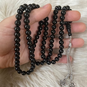 Peut inclure: Un chapelet noir avec un pompon argenté et des embouts décoratifs. Les perles sont rondes et polies, et le pompon est fait de fines chaînes argentées. Les perles sont tenues dans une main.