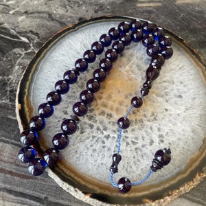 May include: Dark blue prayer beads with a glossy finish. The beads are strung together with a blue cord and feature a decorative tassel. The beads are arranged on a white and brown patterned surface.