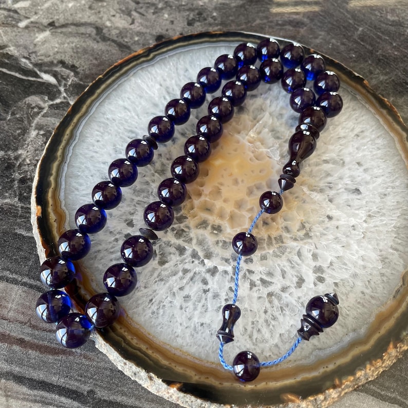 May include: Dark blue prayer beads with a glossy finish. The beads are strung together with a blue cord and feature a decorative tassel. The beads are arranged on a white and brown patterned surface.