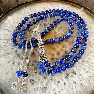 Puede incluir: Un mala de cuentas de oración de lapislázuli azul con un borla de plata y un amuleto de plata. El mala está colocado sobre una rodaja de ágata blanca y gris.