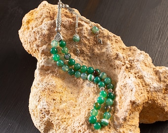 Può includere: Un rosario verde e argento con un pendente a nappa argentato. Le perle sono in agata e hanno un motivo a strisce. Il rosario è appoggiato su una roccia marrone chiaro.