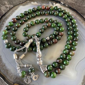 May include: Green and brown gemstone prayer beads with silver accents. The beads are strung on a silver chain with a silver tassel. The beads are arranged in a circle with a silver pendant in the center.