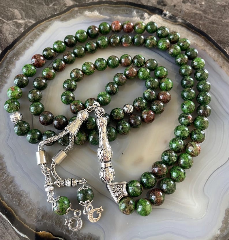 May include: Green and brown gemstone prayer beads with silver accents. The beads are strung on a silver chain with a silver tassel. The beads are arranged in a circle with a silver pendant in the center.