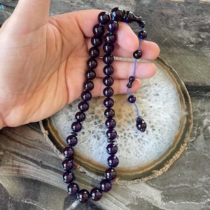 May include: A strand of dark purple prayer beads with a glossy finish. The beads are round and strung together with a blue cord. The tassel and the end piece are also dark purple. The beads are held in a hand.