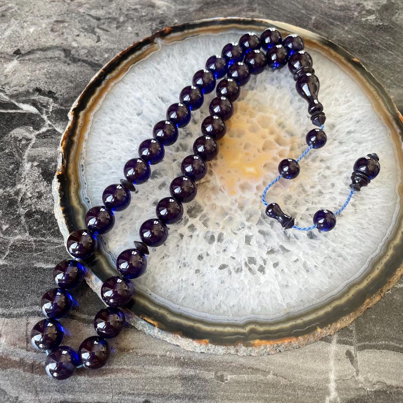 May include: A dark blue beaded prayer rosary. The beads are round and glossy, strung together with a blue cord. The rosary is displayed on a white and brown patterned surface.