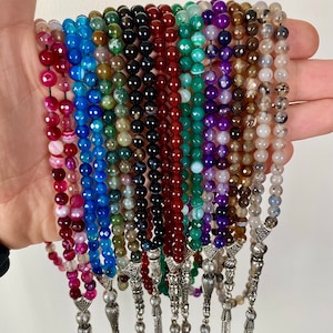 May include: A collection of 18 colorful agate gemstone prayer beads, each with a silver tassel. The beads are strung on a cord and have a variety of colors, including red, blue, green, purple, and brown.