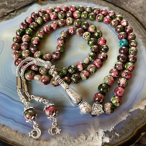 May include: A green and pink prayer beads mala with silver accents. The beads are made of a speckled stone and the tassel is silver with a decorative design.