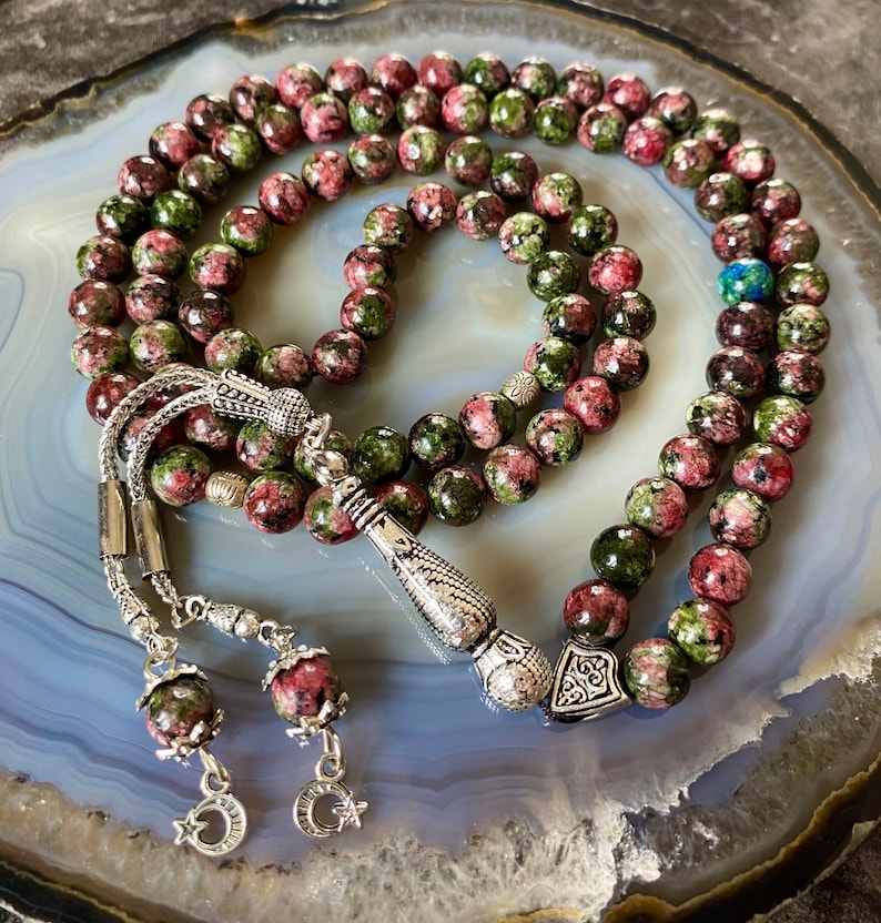 May include: A green and pink prayer beads mala with silver accents. The beads are made of a speckled stone and the tassel is silver with a decorative design.