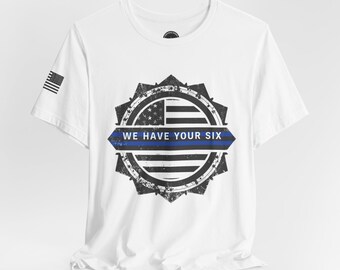 May include: White t-shirt with a distressed design featuring an American flag and a blue line, and the text "WE HAVE YOUR SIX". The shirt also has a small American flag on the sleeve.
