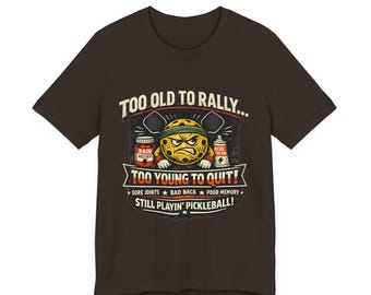 May include: A dark brown t-shirt with a humorous graphic. The design features a yellow pickleball character with an angry face, and text that reads "Too Old to Rally... Too Young to Quit! Still Playin' Pickleball!" Includes pain relief and ache cream graphics.