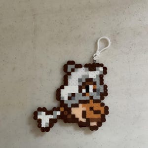 May include: A pixelated keychain featuring a brown, white, and tan character. The character has a bone-like structure and a small white loop for attachment. The design is made of small, colorful beads.