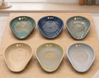 May include: A set of six ceramic spoon rests arranged in two rows. Each spoon rest features a teardrop shape, a smooth glazed finish in various muted colors, and a textured circular pattern in the center with three small holes at the top.
