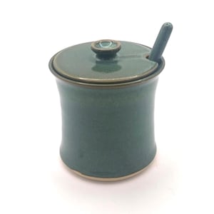 May include: A teal ceramic sugar bowl with a matching lid and a small spoon. The bowl has a rounded shape with a slightly flared base. The lid has a small knob for easy removal. The spoon rests in a notch on the lid.
