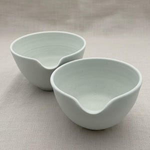 May include: Two off-white ceramic bowls with a speckled finish and a smooth matte texture. Each bowl features a distinct pouring spout integrated into the rounded rim, creating a functional design for kitchen use with a deep, open basin.