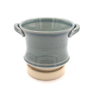 Pu&ograve; includere: Un vaso in ceramica con una finitura lucida color blu-grigio e due piccole maniglie laterali. Il contenitore presenta un bordo svasato e poggia su una base circolare separata, non smaltata e di colore beige, che offre stabilit&agrave;.
