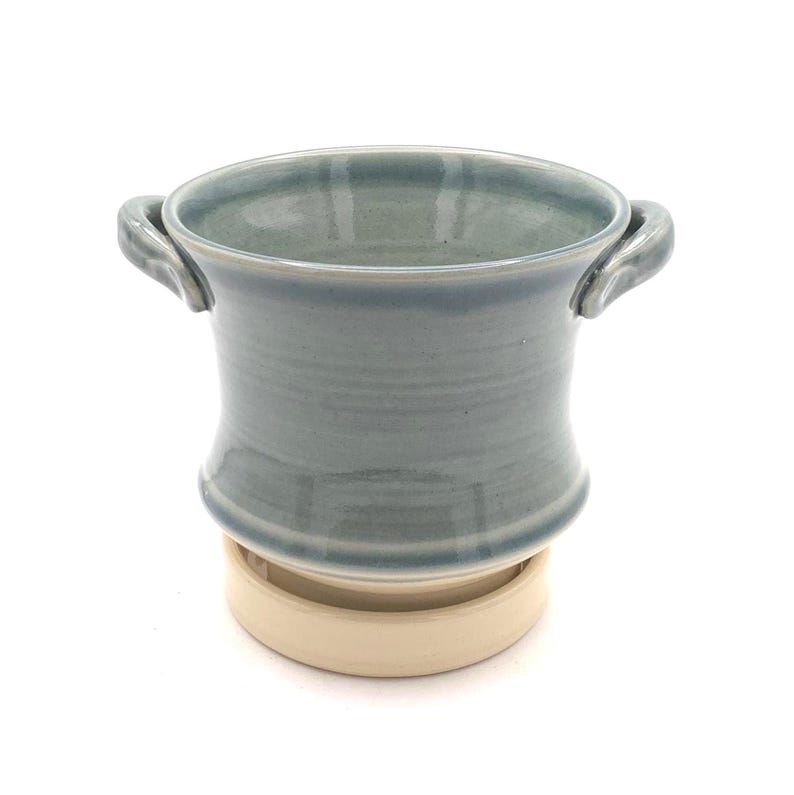 Pu&ograve; includere: Un vaso in ceramica con una finitura lucida color blu-grigio e due piccole maniglie laterali. Il contenitore presenta un bordo svasato e poggia su una base circolare separata, non smaltata e di colore beige, che offre stabilit&agrave;.