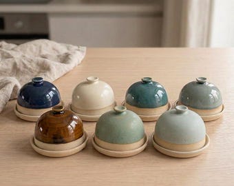 May include: A collection of seven small ceramic butter dishes with dome-shaped lids. Each dish features a two-tone design with a glazed top in various colors including navy, cream, teal, brown, and sage, paired with a natural matte base.