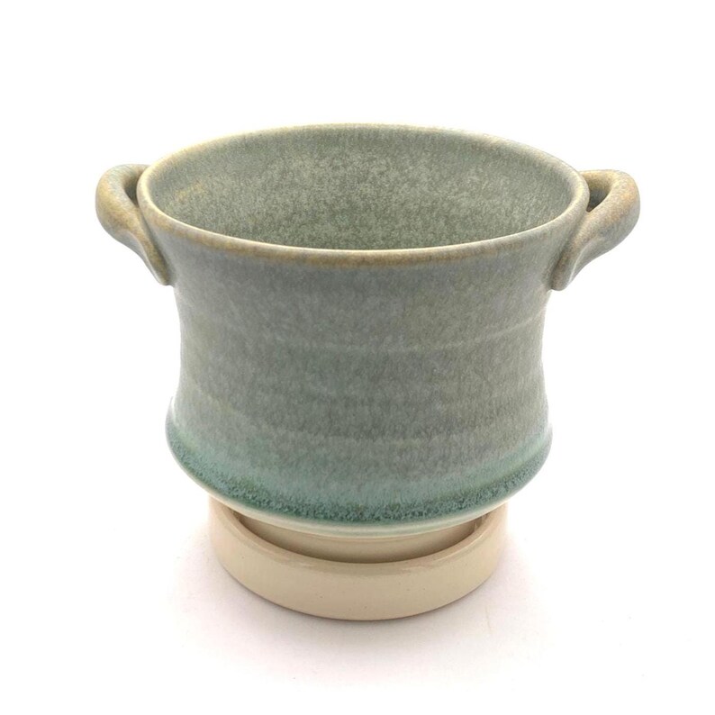 Pu&ograve; includere: Una ciotola in ceramica con due piccole maniglie laterali, caratterizzata da uno smalto verde salvia maculato che sfuma in una tonalit&agrave; verde acqua pi&ugrave; scura verso la base. La ciotola poggia su una base circolare liscia color bianco sporco.