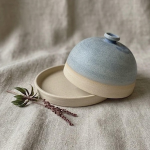 May include: A ceramic butter dish consisting of a flat, circular, beige base and a dome-shaped lid with a speckled, light blue glaze. The lid features a small, circular knob at the top. A small sprig of dried green leaves lies beside the dish.
