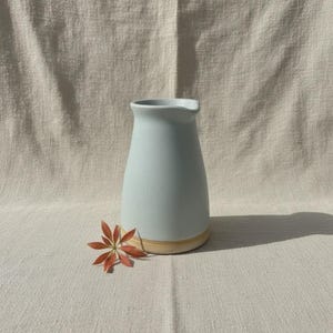 Pode incluir: Um jarro de cerâmica azul-claro com um acabamento mate suave e uma textura subtilmente salpicada. A base do jarro apresenta um rebordo natural não vidrado de cor castanha. Um pequeno e delicado ramo de planta vermelho-alaranjado repousa na base.