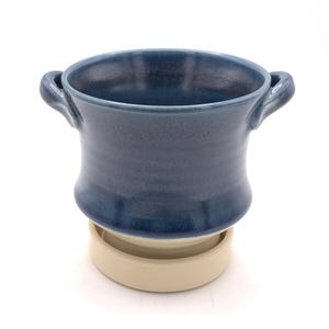 Pu&ograve; includere: Un vaso da fiori in ceramica con smalto blu lucido e due manici laterali, poggiato su un piattino circolare beige non smaltato. Il contenitore presenta un bordo largo e svasato e un corpo liscio e arrotondato che si restringe verso la base.
