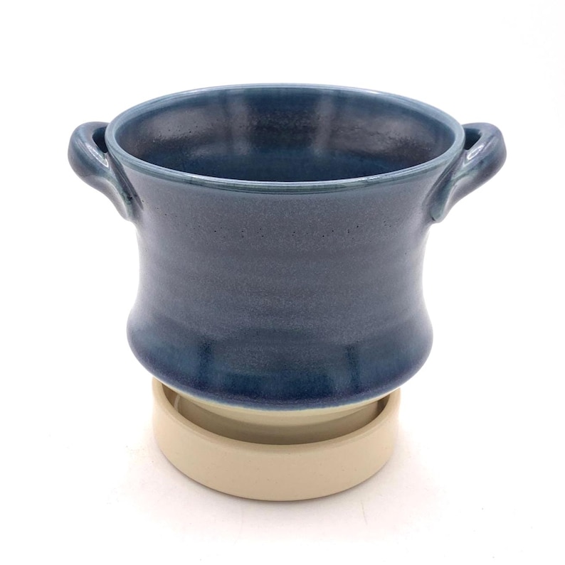 Pu&ograve; includere: Un vaso da fiori in ceramica con smalto blu lucido e due manici laterali, poggiato su un piattino circolare beige non smaltato. Il contenitore presenta un bordo largo e svasato e un corpo liscio e arrotondato che si restringe verso la base.