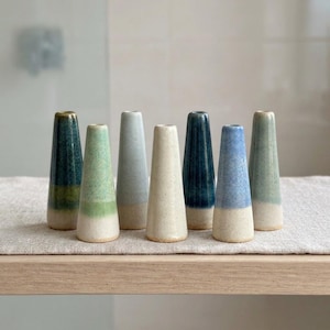 May include: A collection of seven conical ceramic bud vases arranged in a row. Each vase features a unique glaze finish in varying shades of blue, green, and cream, with a smooth, tapered silhouette and a small circular opening at the top.
