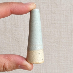May include: A conical ceramic object with a speckled light blue glaze on the top portion and a textured, unglazed beige base. The item is held vertically, showcasing its tapered shape and distinct two-tone colour transition.