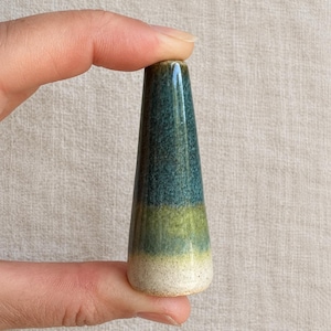 May include: A conical ceramic object featuring a smooth, glazed finish with a colour gradient transitioning from a deep teal at the top to a muted olive green in the middle and a speckled off-white base at the bottom.
