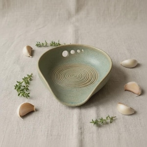 Puede incluir: Un plato rallador de cerámica color verde salvia con un centro circular texturizado para rallar ajo o jengibre. El plato presenta un borde liso, un pequeño pico vertedor y cuatro orificios circulares graduados en el borde superior, rodeado de dientes de ajo y ramitas de tomillo.