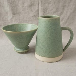 May include: A ceramic pour-over coffee dripper and a matching pitcher with a handle. Both items feature a speckled sage green glaze on the upper sections and a solid off-white base. The dripper has a wide, conical shape, while the pitcher is cylindrical.