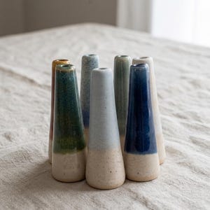 May include: A collection of seven ceramic bud vases arranged in a cluster. Each vase features a tapered, conical shape with a narrow opening at the top. The vases display various glaze finishes, including solid deep blue, light blue, green, and beige.