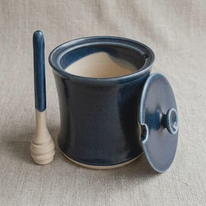 Puede incluir: Un tarro de miel cilíndrico de cerámica con un exterior esmaltado en azul profundo y un interior de color crema. El tarro incluye una tapa circular a juego con un pequeño pomo y una muesca lateral, junto a un largo cucharón de miel bicolor.