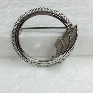 May include: A silver brooch with a circular design and a decorative, abstract leaf-like shape.