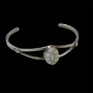 May include: A silver cuff bracelet with an oval, inlaid mother-of-pearl centerpiece. The bracelet has a simple, open design with a curved band and decorative accents. The centerpiece is made of white and cream-colored pieces.
