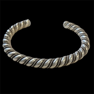 May include: Silver-toned cuff bracelet with a twisted rope design. The bracelet has a thick, rounded form with a continuous pattern of interwoven strands. The open ends allow for easy wear. The background is solid black.