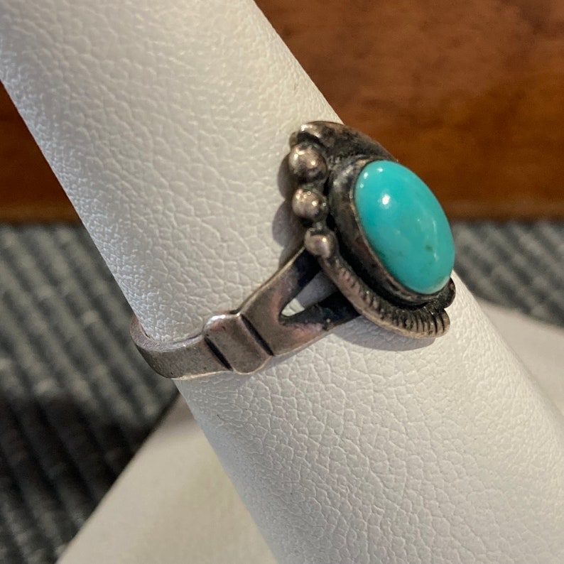K&ouml;nnte beinhalten: Ein silberner Ring mit einem T&uuml;rkis-Stein, der in einem dekorativen Design gefasst ist. Der Ring befindet sich auf einem wei&szlig;en Pr&auml;sentationsst&auml;nder.
