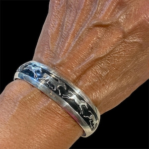May include: A silver cuff bracelet with a black inlay featuring a repeating pattern of running horses. The bracelet is worn on a wrist, showcasing its design and craftsmanship. The bracelet has a polished finish.