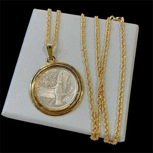 Może przedstawiać: Złoty naszyjnik z okrągłą zawieszką z motywem srebrnej monety. Moneta przedstawia orła z rozpostartymi skrzydłami i napisem "SILVER ISSUE". Łańcuszek jest w delikatnym stylu sznurkowym, a zawieszka jest oprawiona w złoto.