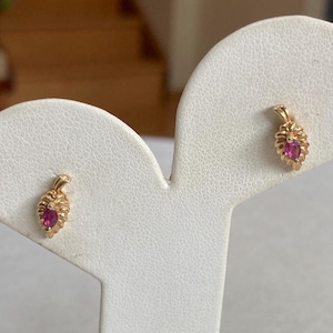 May include: A pair of gold earrings with a pink gemstone in the center of each earring. The earrings are on a white display stand.
