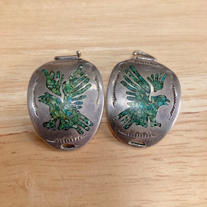 Op de afbeelding: Een paar zilveren manchetknopen met turquoise inleg, die een gestileerd vogelontwerp voorstellen.